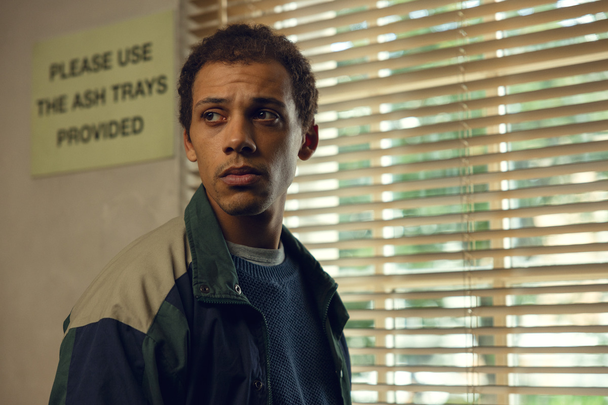 A man in a casual jacket stands indoors near a window with blinds, looking to the side. Behind him is a sign on the wall that reads "Please use the ash trays provided." The setting appears to be a public or communal space.