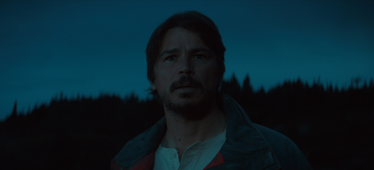 A man with facial hair stands outdoors at dusk, looking ahead with a concerned expression, silhouetted trees and a dark blue evening sky in the background.