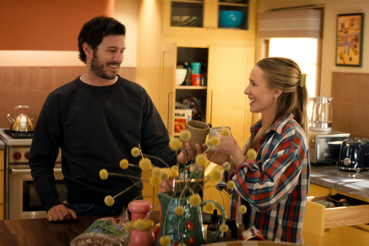 Homem e mulher sorriem e conversam com canecas nas mãos, em uma cozinha aconchegante e bem iluminada, com armários de madeira, plantas decorativas e pão sobre o balcão.