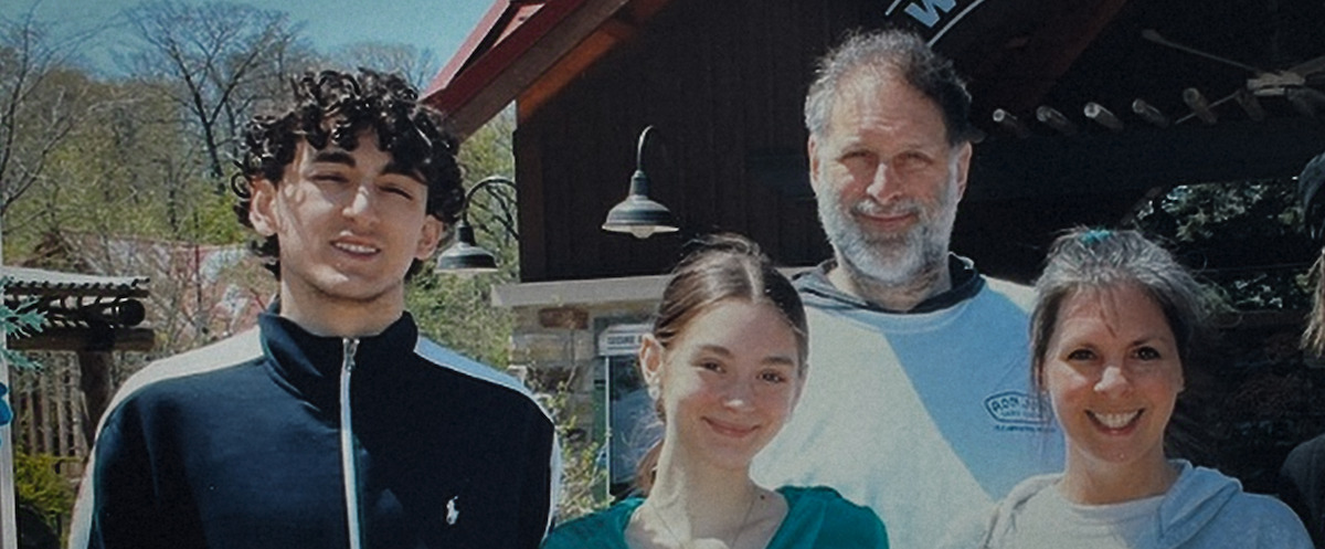 Four people standing together outdoors, smiling in front of a rustic building with a sloped roof and outdoor lighting, trees and blue sky in the background, casual daytime setting.Four people standing together outdoors, smiling in front of a rustic building with a sloped roof and outdoor lighting, trees and blue sky in the background, casual daytime setting.