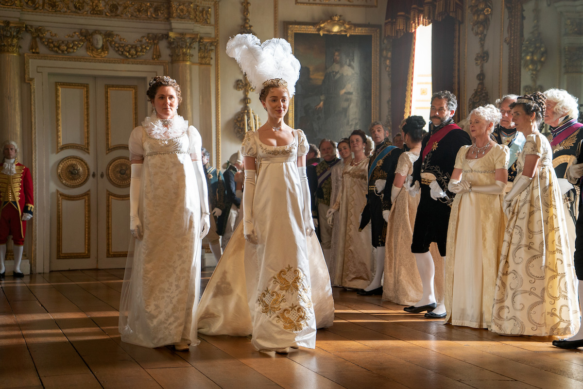 Two women in elegant white Regency-era gowns walk into a lavishly decorated ballroom, watched by formally dressed guests, under ornate gold-trimmed walls and paintings, suggesting a high-society historical event or royal gathering.