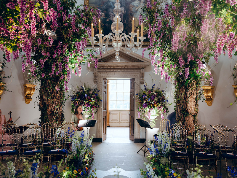 A room filled with flowers along an aisle, along the walls, and hanging from the ceiling in 'The Event of the Season: A Bridgerton Wedding'