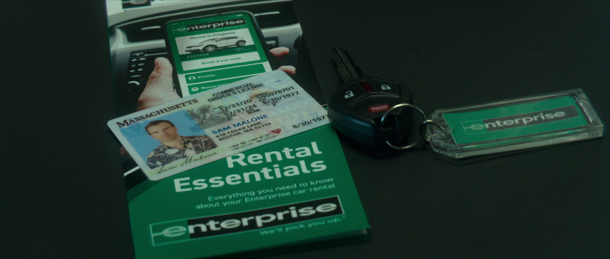 A Massachusetts driver’s license that reads Sam Malone sits on top of an Enterprise car rental pamphlet and beside a car key.