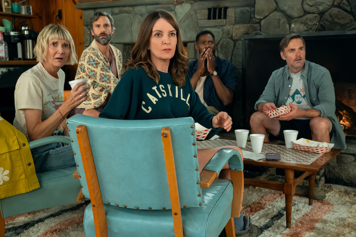 Five people sit in a cozy living room with a stone fireplace, looking surprised at something off-camera. There are snacks and cups on the table, and the group appears to be in mid-conversation or reaction.