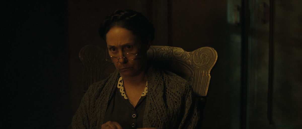An older woman with glasses and braided hair sits in a carved wooden chair, looking sternly ahead in a dimly lit room.