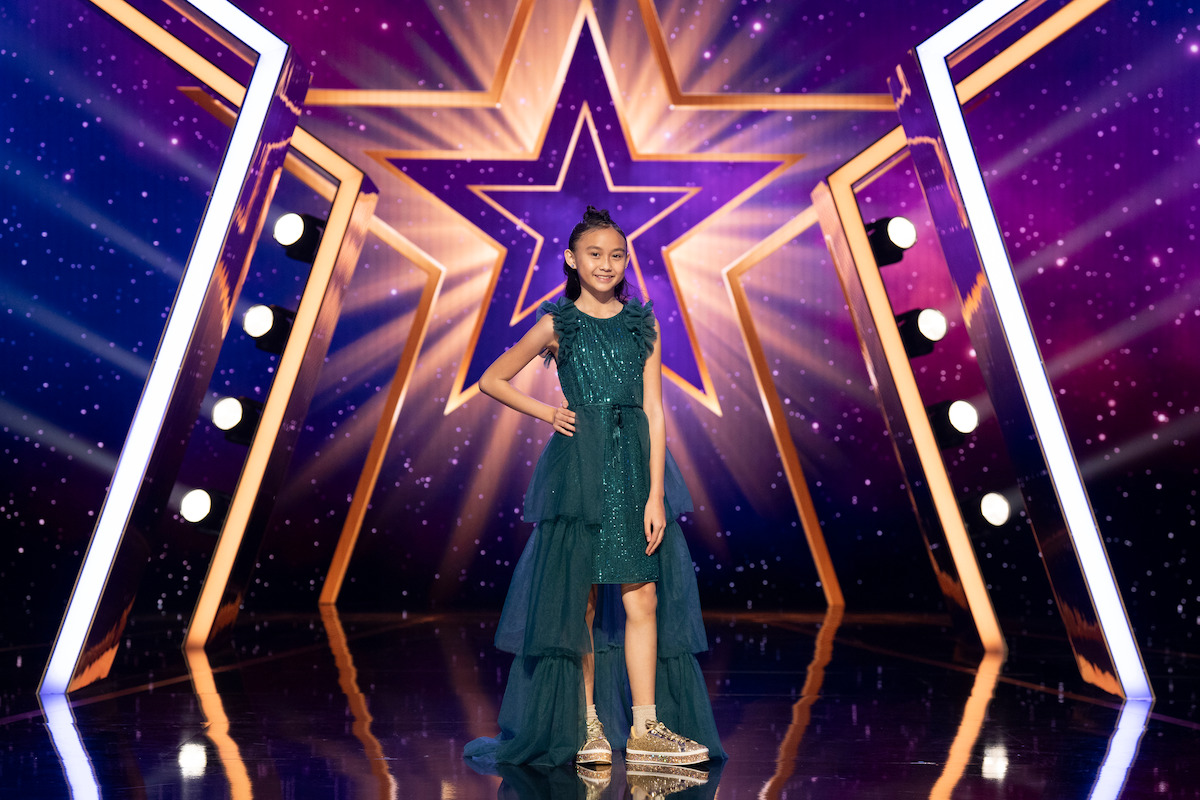 Madison Lauren in a green dress poses confidently on a shiny stage with dramatic lighting, a large illuminated star backdrop, and colorful purple and gold decorations, suggesting a talent show or competition setting.