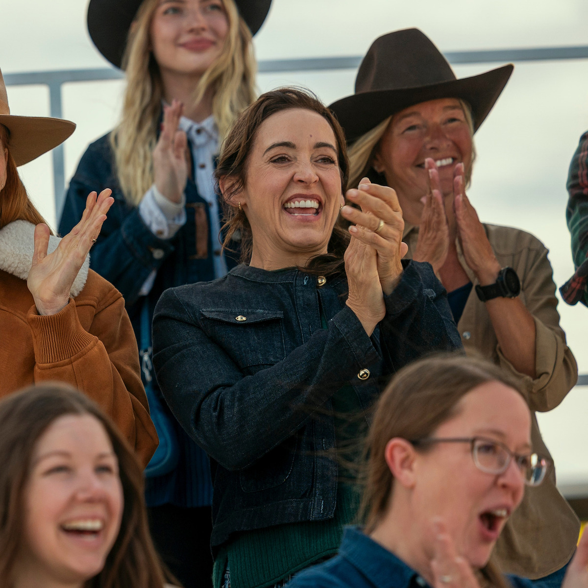 Group of women outdoors in casual clothing and hats, smiling and clapping in a stadium or bleacher setting, suggesting they are cheering or celebrating at an event.