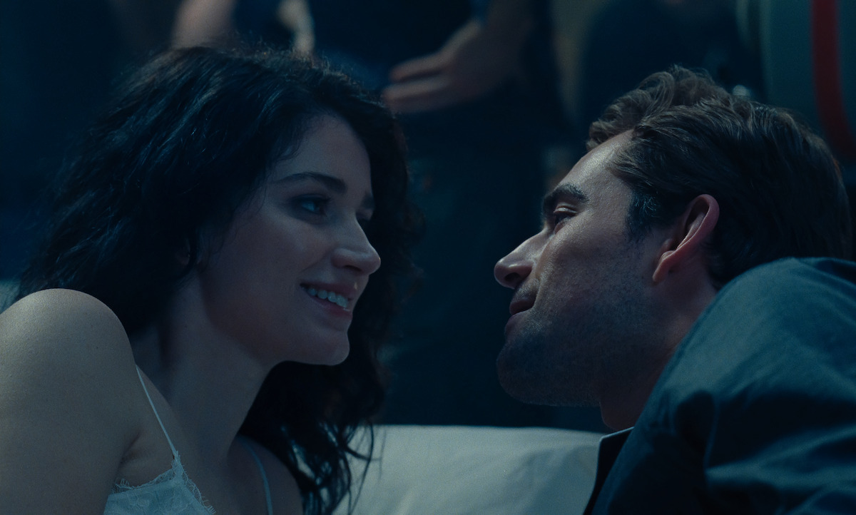 A woman and man lie close together on a bed, gazing at each other and smiling softly in a dimly lit, intimate setting, with a relaxed and romantic atmosphere.