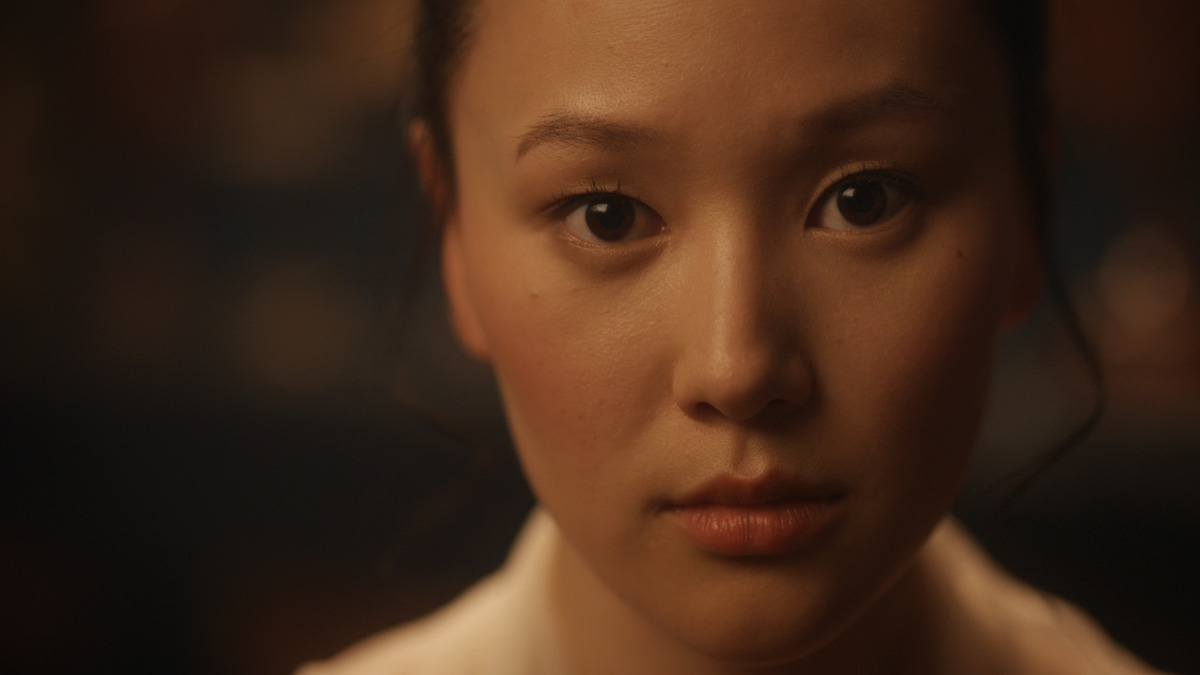 A young woman with dark hair looks directly at the camera with a neutral expression, softly lit in a warm indoor environment with a blurred background.
