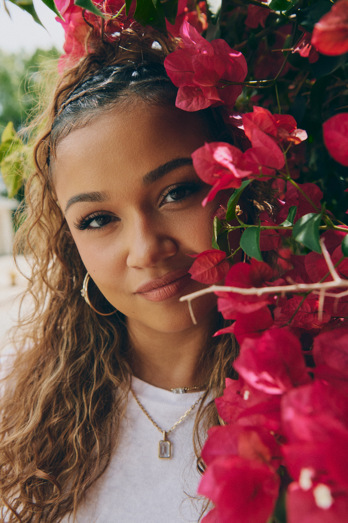 Young woman with curly hair and gold jewelry poses outdoors beside vibrant pink bougainvillea flowers, soft natural light highlighting her face and the lush green foliage around her.