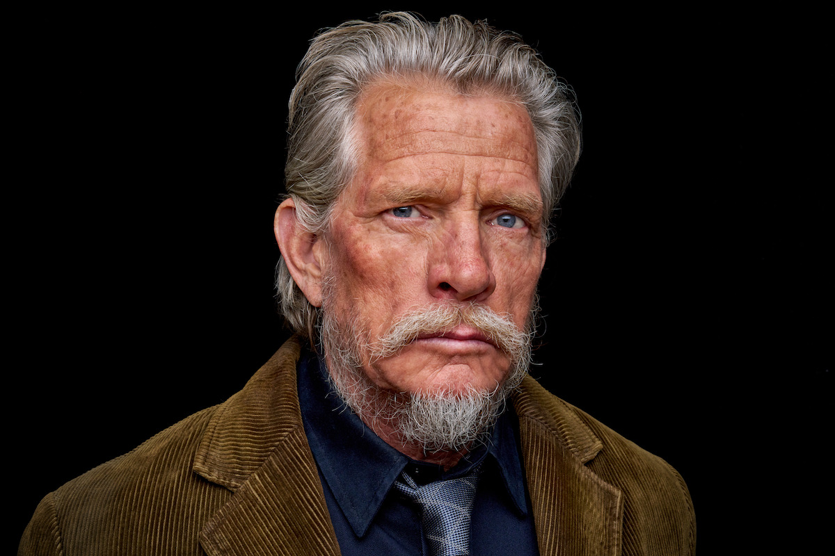 Older man with gray hair and beard, wearing a corduroy jacket, shirt, and tie, stands against a plain black background, displaying a serious and thoughtful expression in a studio portrait setting.