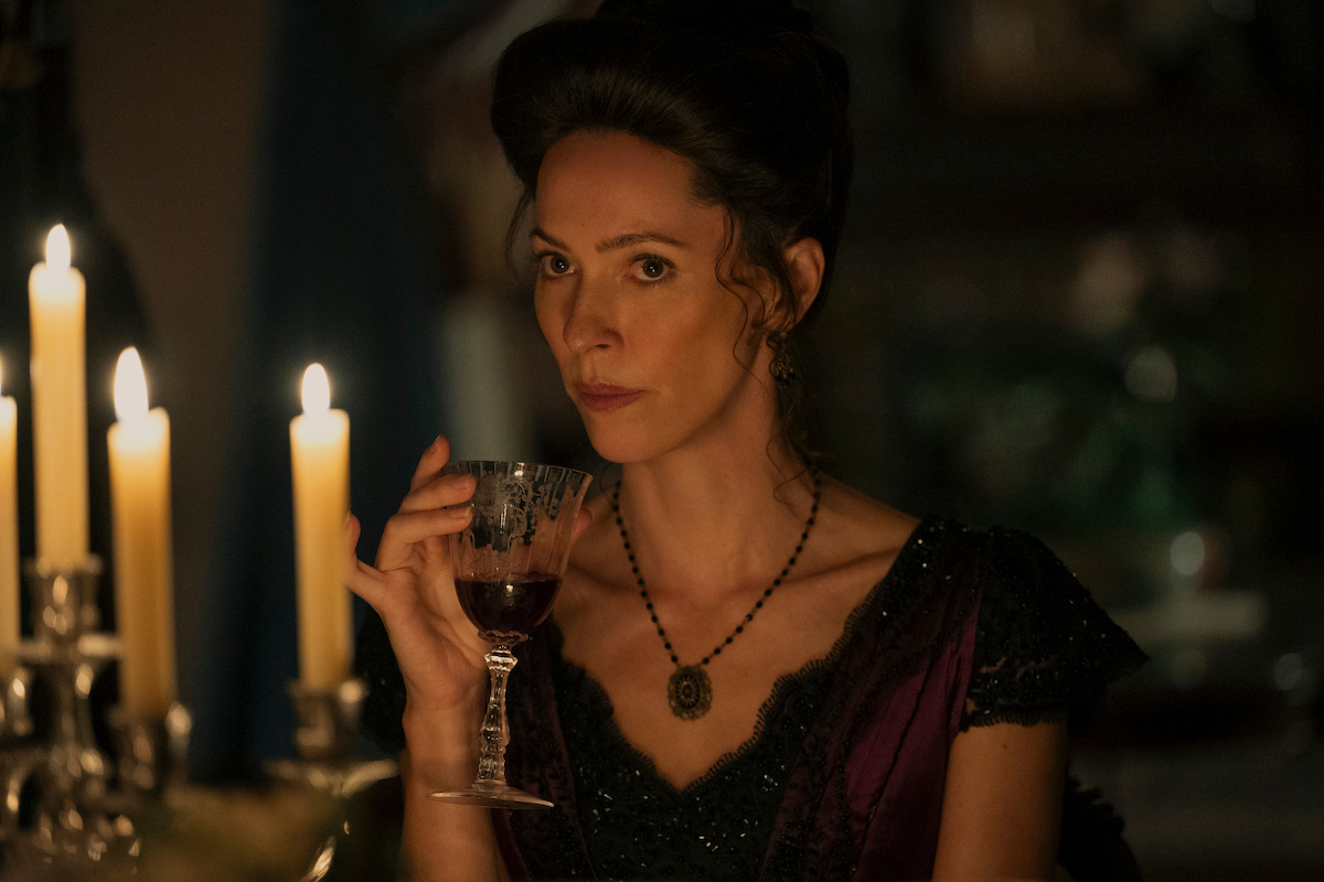 Woman in period costume sits at a candlelit table, holding a glass of red wine, with a serious expression in a dimly lit, elegant room.