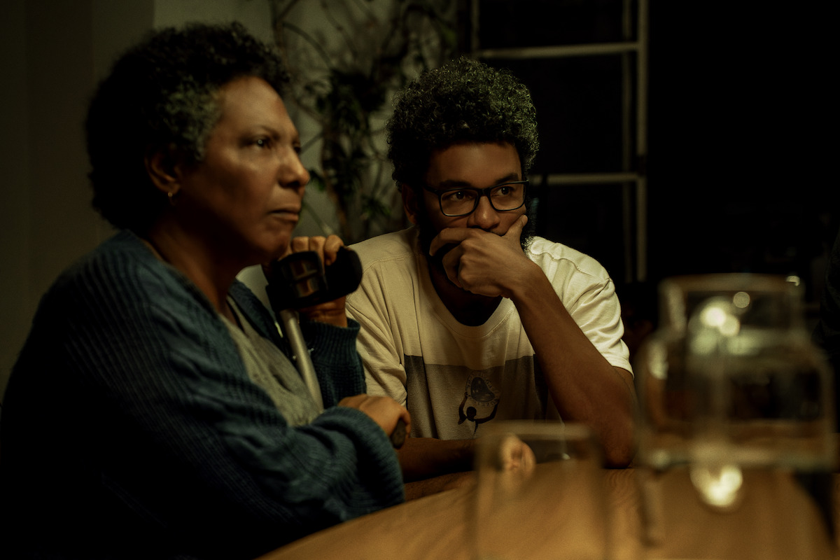 Two people sitting at a wooden table in a dimly lit room; one holds a phone, both look serious and thoughtful. There are glasses on the table and a plant in the background near a window.
