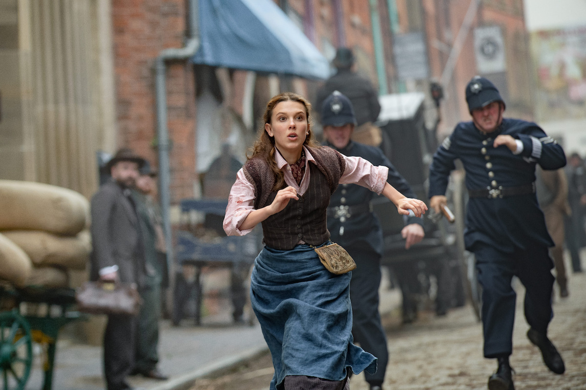 A woman in period clothing runs down an alley while a group of policemen chase her.