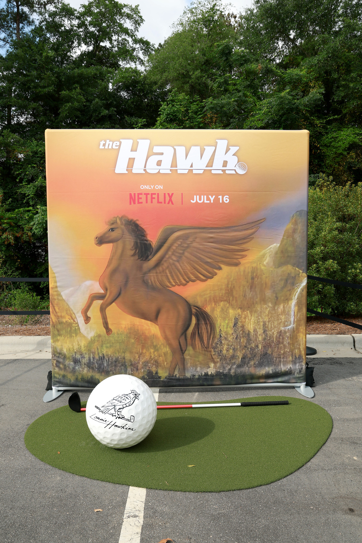 Promotional display for "the Hawk" on Netflix shows a winged horse on a nature backdrop, with a giant golf ball and small green mat in a parking lot, surrounded by trees in the background.