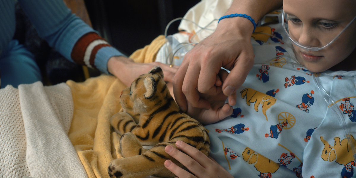 A child in a hospital bed wearing an oxygen tube holds a plush tiger, comforted by the hand of an adult.