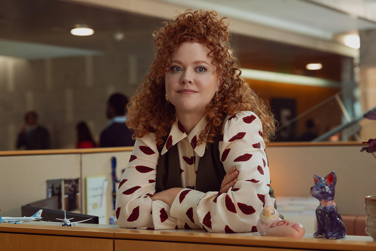 Woman with curly red hair in a modern office, leaning on a desk decorated with figurines, model planes, and papers. The environment includes other people in the background and a contemporary indoor setting.