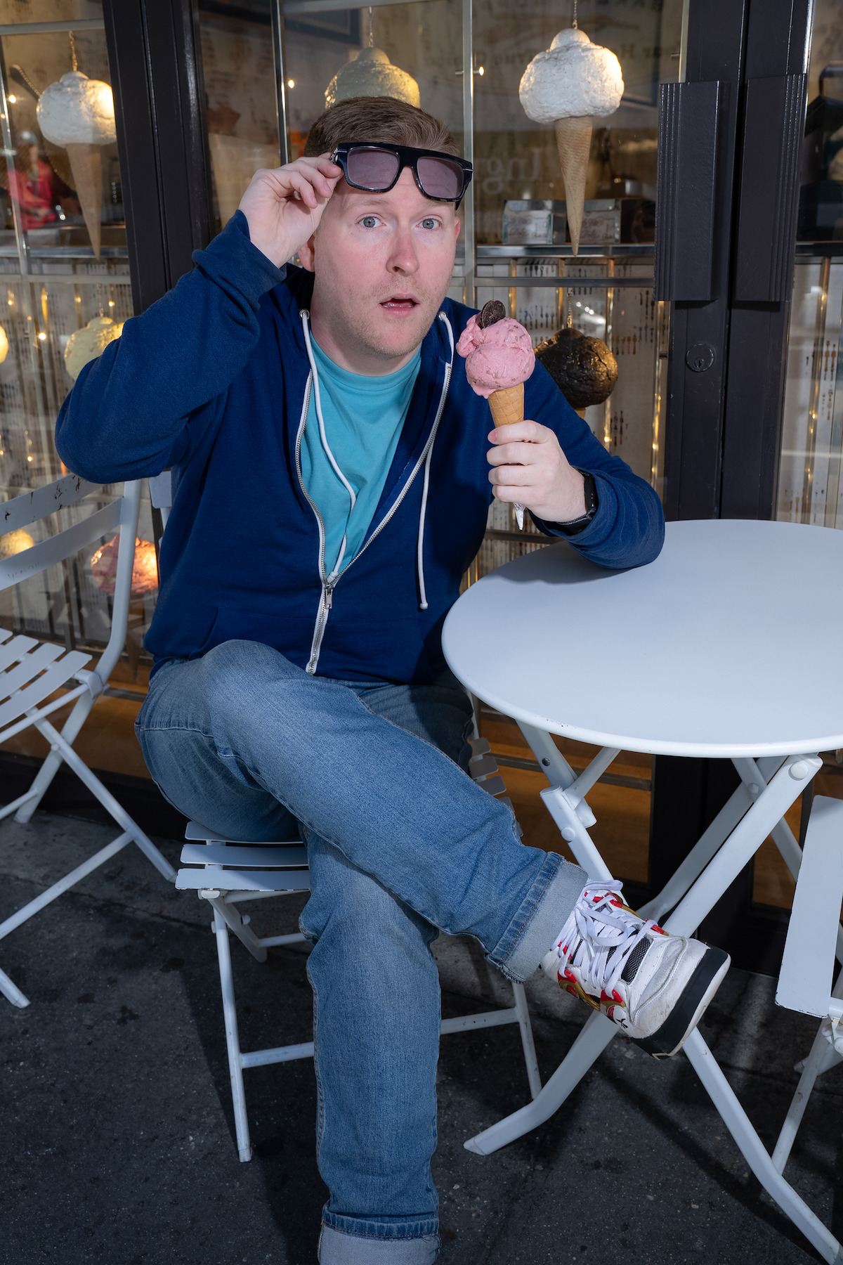 Man in casual clothes sitting at an outdoor café table, holding a cone of pink ice cream and lifting his glasses with a surprised expression, with shop window and lights in the background.