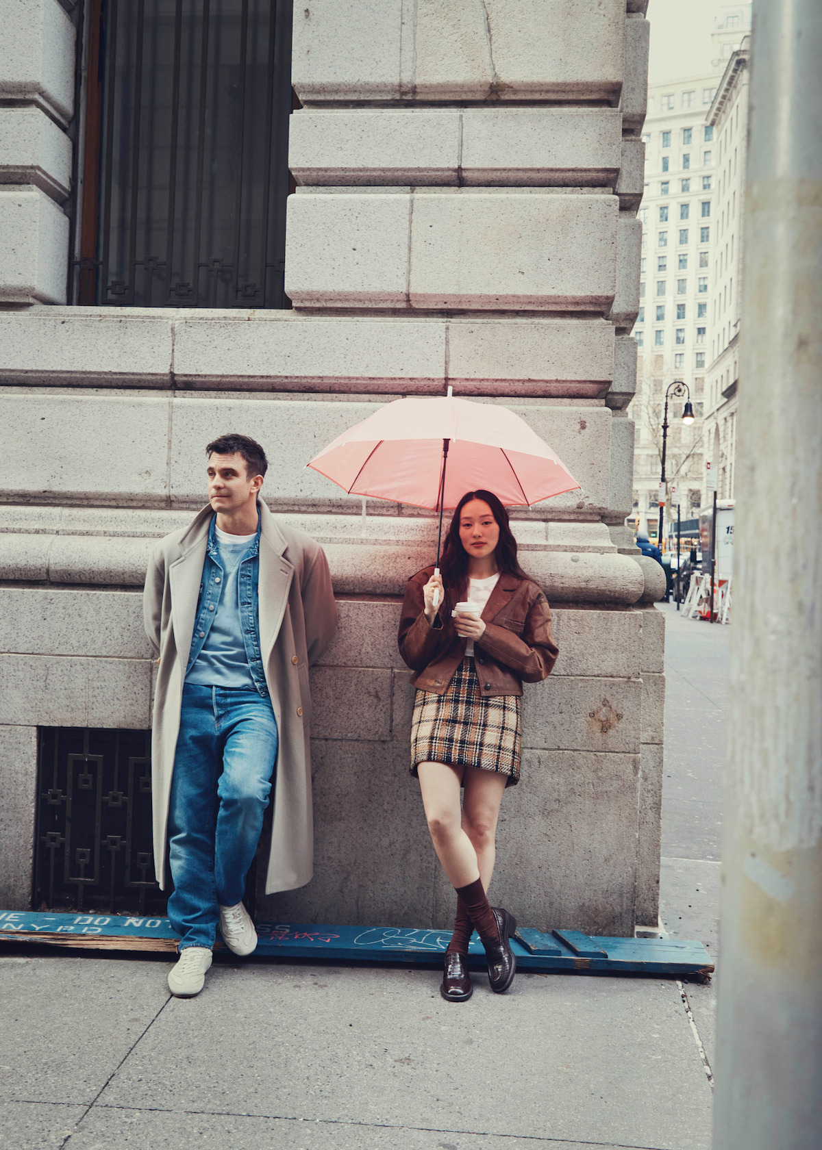 A man in a long coat and a woman holding a pink umbrella and a drink stand against a stone building on a city street, dressed in stylish, casual clothes on a cloudy day.