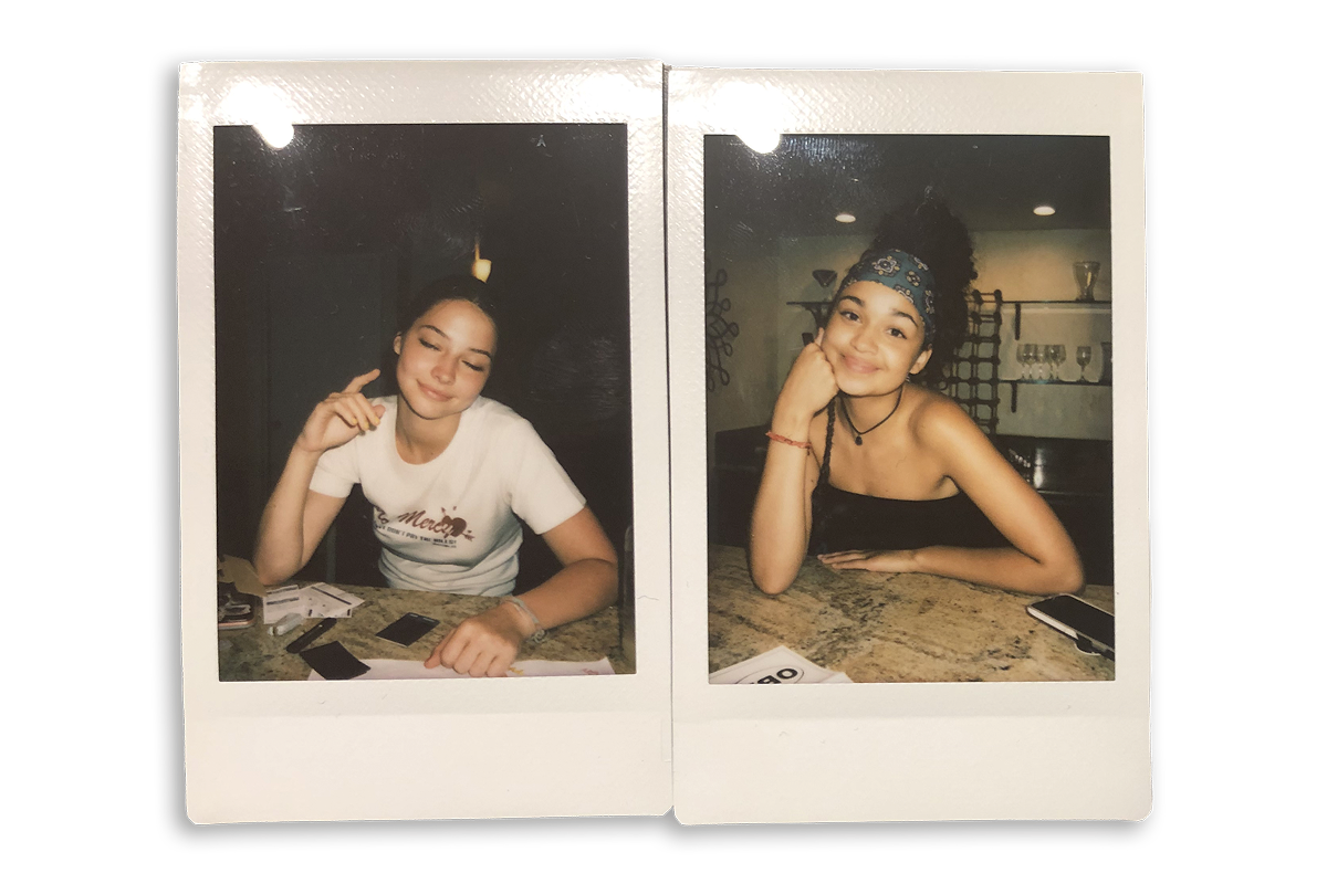 Two young women posing separately at a kitchen counter, each smiling and resting their heads on their hands. The environment appears casual, with kitchen decor and various items on the counter in a warmly-lit indoor setting.