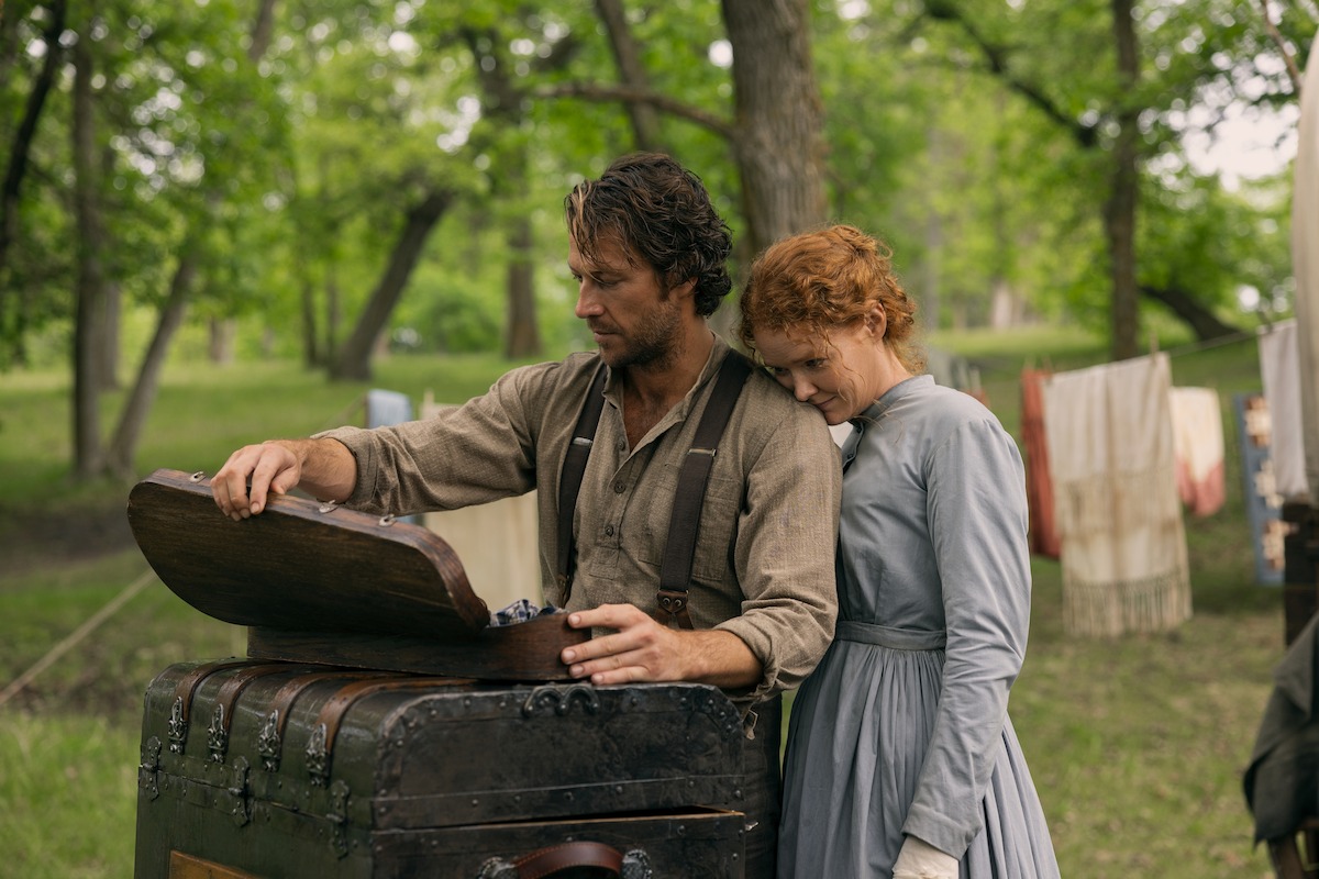 A man opens an old chest while a woman leans on his shoulder. They are outdoors in a wooded area with laundry hanging in the background, suggesting a historical or pioneer setting.