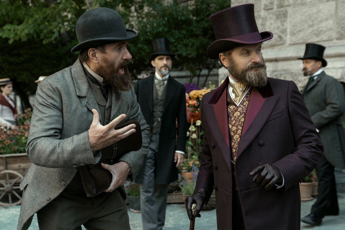 Two men in Victorian-era attire converse outdoors; one gestures animatedly while the other listens holding a cane. Background shows other well-dressed men, potted flowers, and stone building, creating a historical setting.