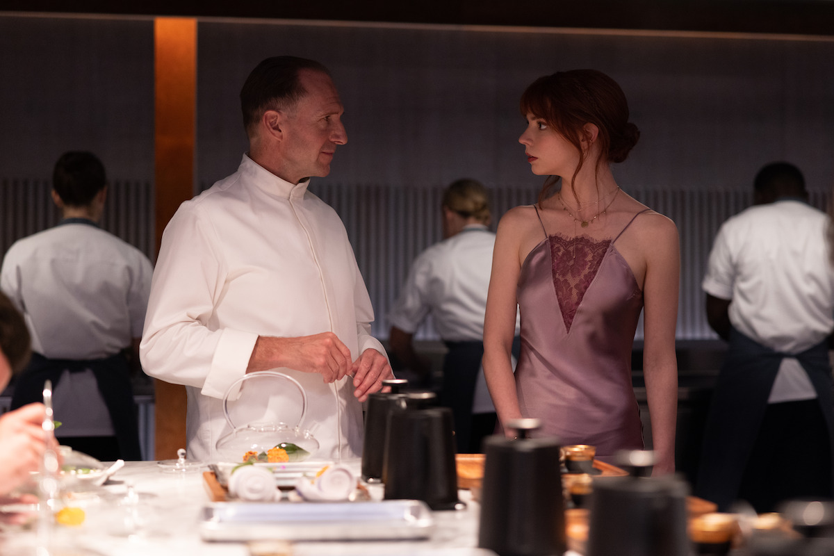 A chef in a white uniform talks to a woman in a purple dress at a modern restaurant counter, with kitchen staff working in the background. The atmosphere is upscale and focused.