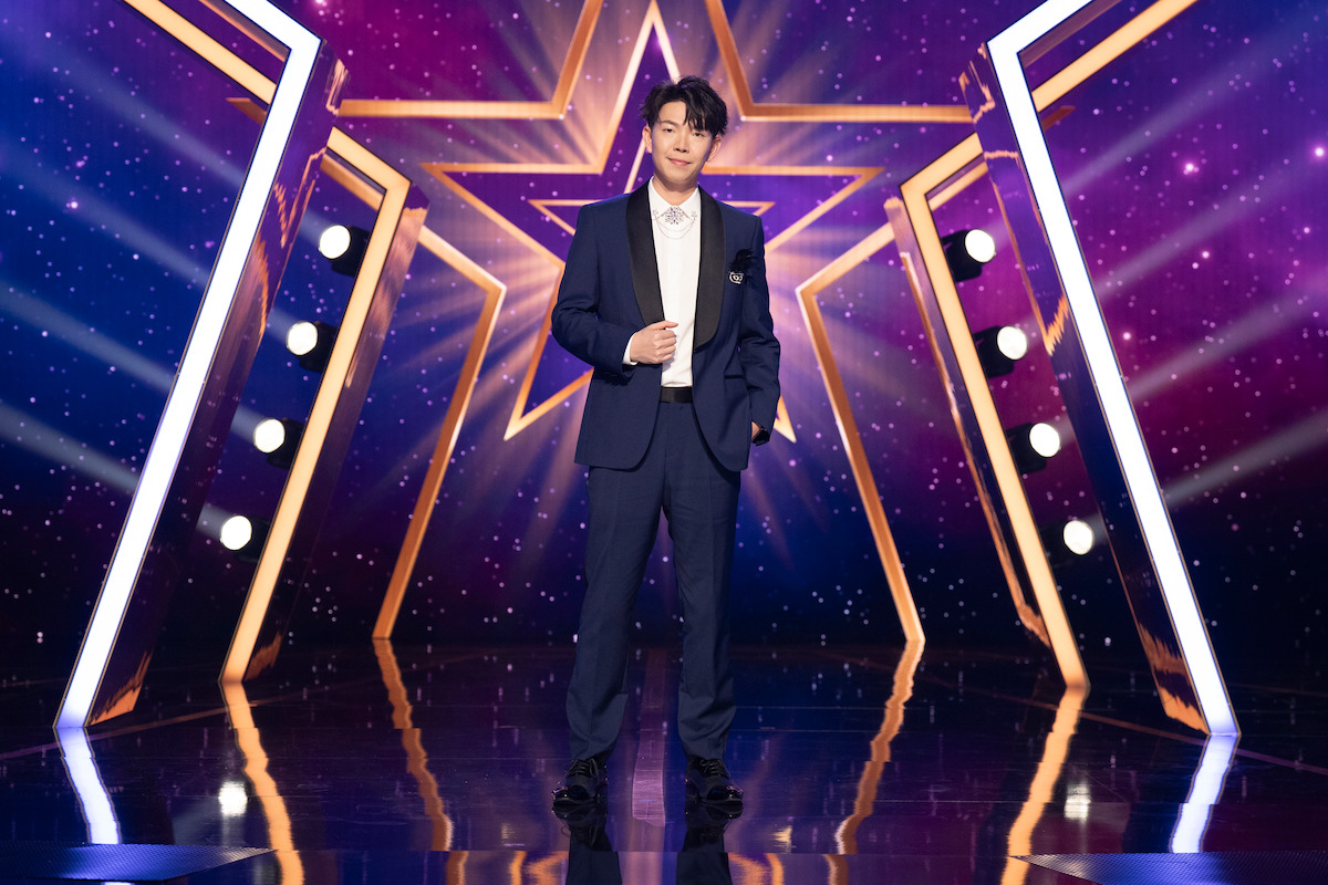 Johnny Wu stands on a shiny stage with star-shaped golden lights and a purple, starry background, creating a glamorous, show-like atmosphere.