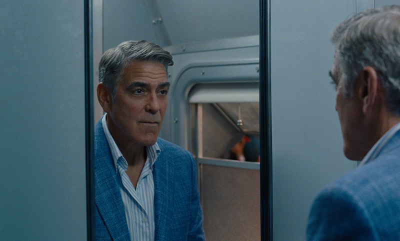 Jay Kelly (George Clooney) wears a suit and stares into a mirror in the restroom of an Italian passenger train.