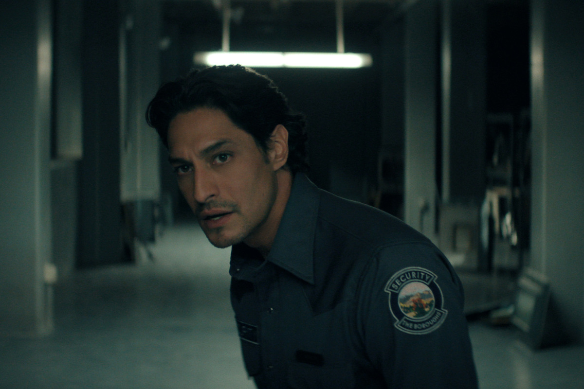 Carlos Miranda as Paz Navarro, a security guard in uniform stands alert in a dimly lit hallway, with gray walls and scattered objects in the background, conveying a tense or suspenseful atmosphere in an indoor setting.