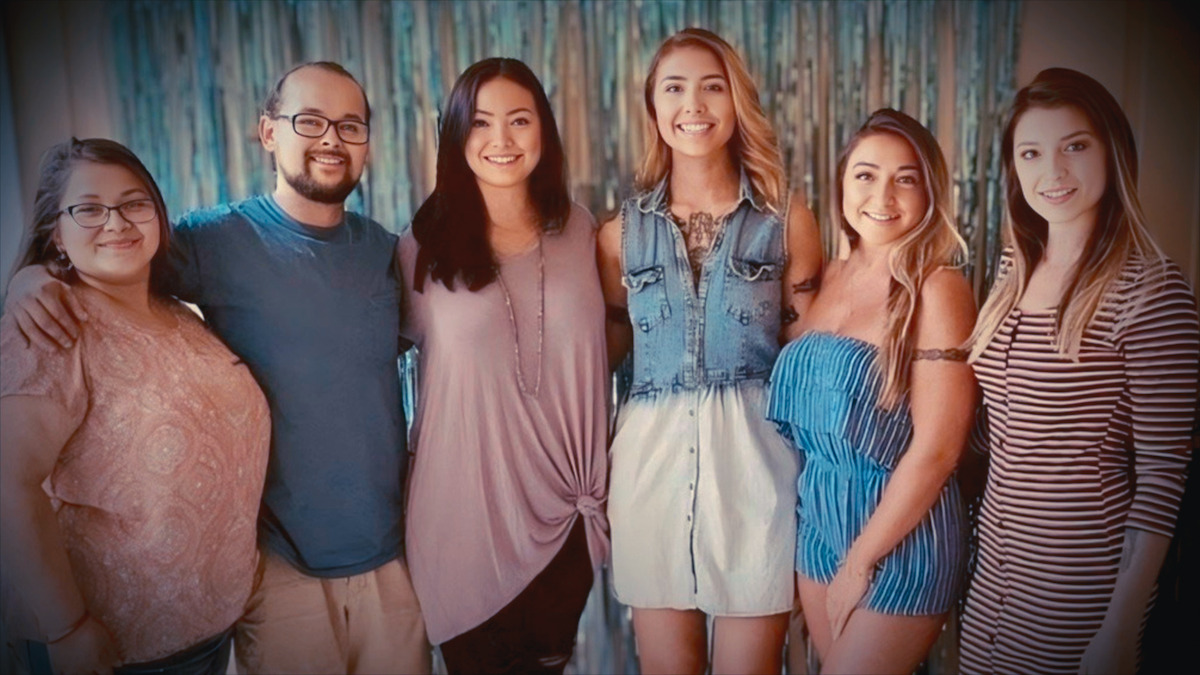 Six smiling young adults standing together indoors in front of a blue and white striped backdrop, posing for a group photo in casual clothing.