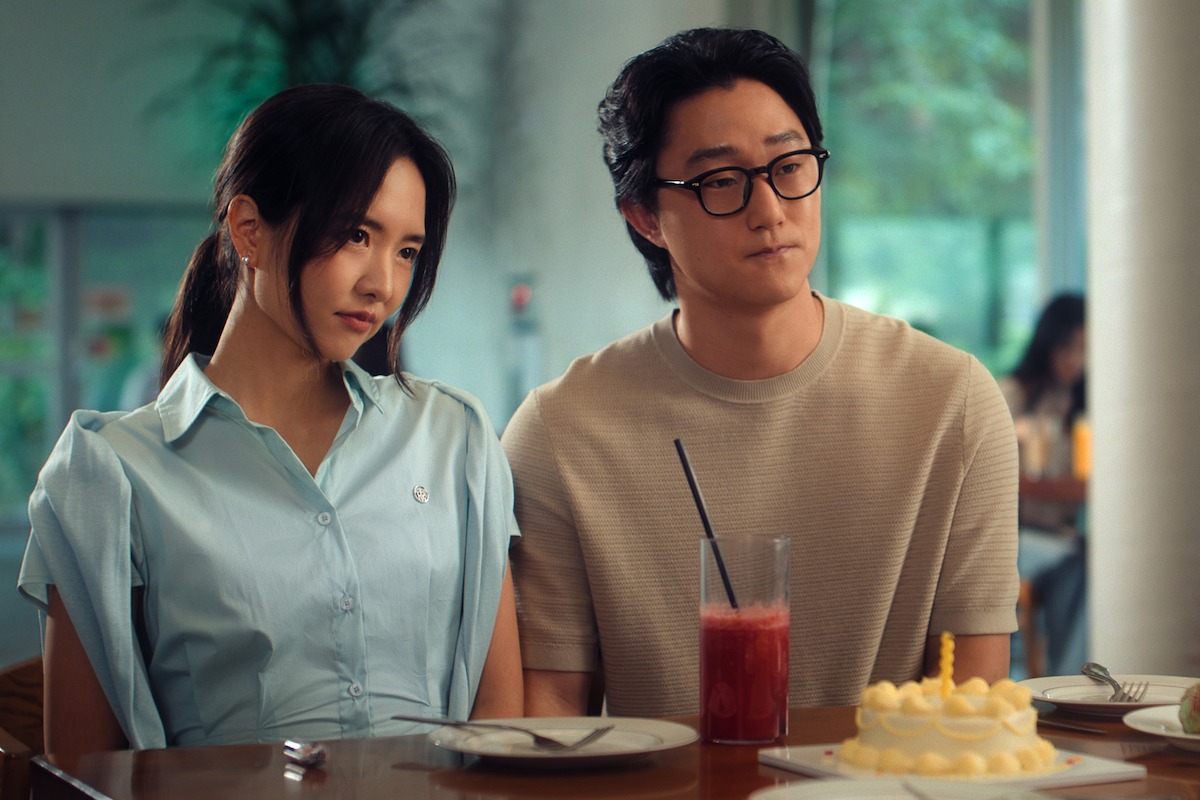 Two people sit together at a table in a bright, modern café, looking pensively at a birthday cake with a candle and a glass of red drink, suggesting a contemplative mood during a small celebration.
