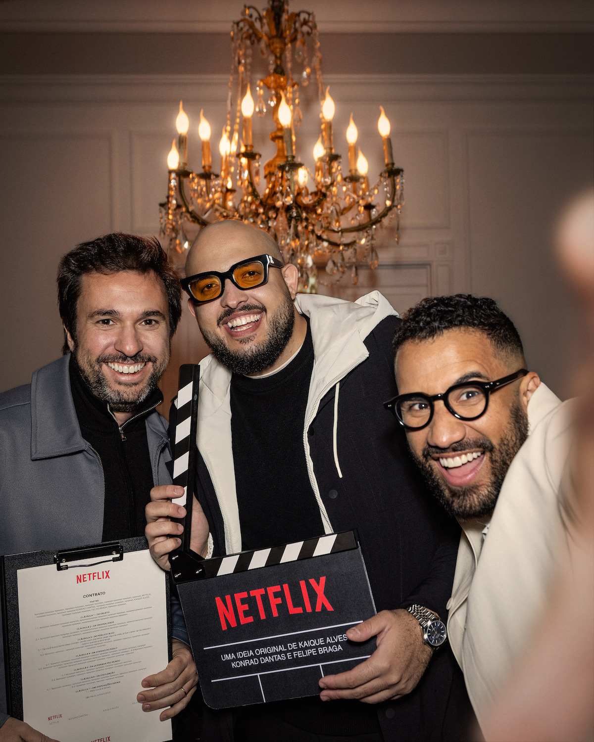 Três homens posam lado a lado enquanto seguram uma claquete na qual se lê “Netflix”