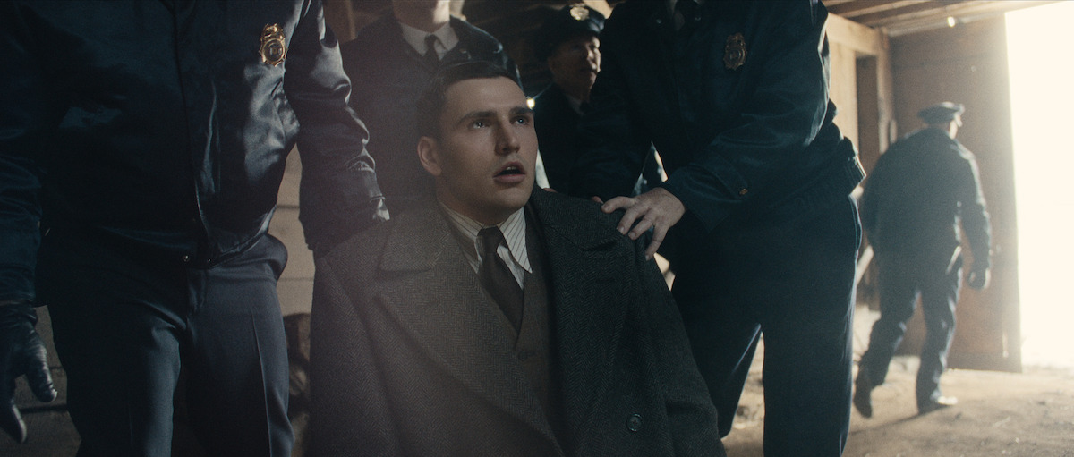 A young man in a suit coat is held by police officers, looking distressed as he is led out of a dimly lit building toward a bright exit.