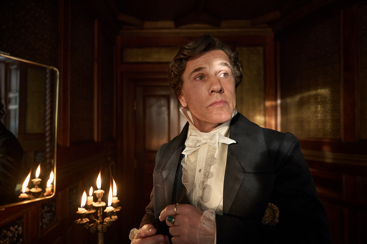 A man in historical attire stands in a dimly lit, ornately decorated room beside a lit candelabrum, looking contemplative and serious, evoking a dramatic, vintage atmosphere.