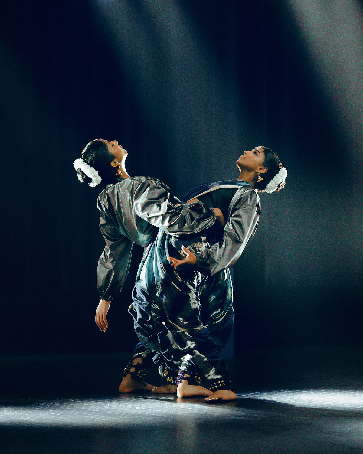 Two dancers in traditional attire perform an expressive pose on a dimly lit stage, with dramatic spotlights highlighting their coordinated movement and elegant costumes against a dark background.