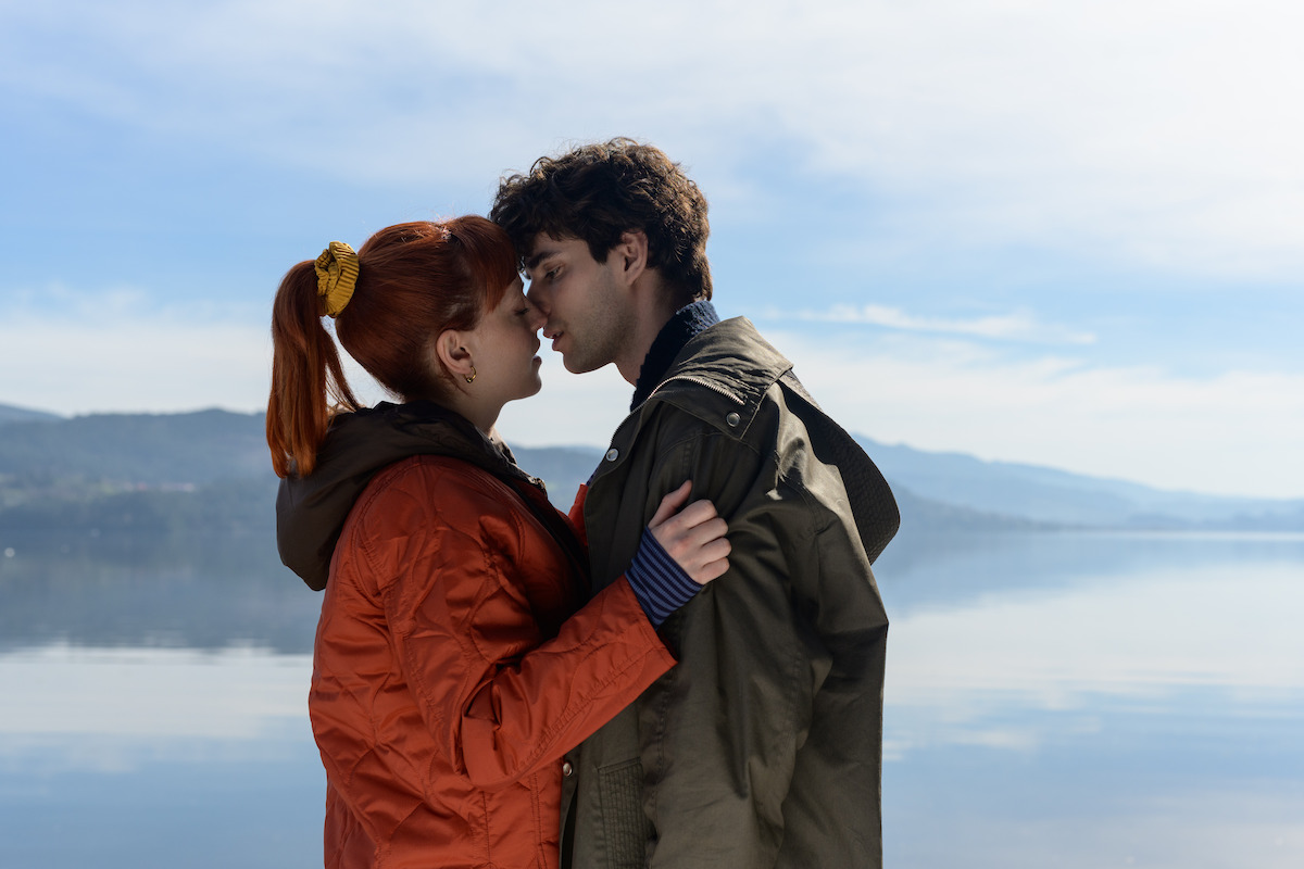 In an image from the series ‘The Gardener,’ Catalina Sopelana as Violeta and Álvaro Rico as Elmer embrace while wearing winter jackets with a lake and low mountains in the background.