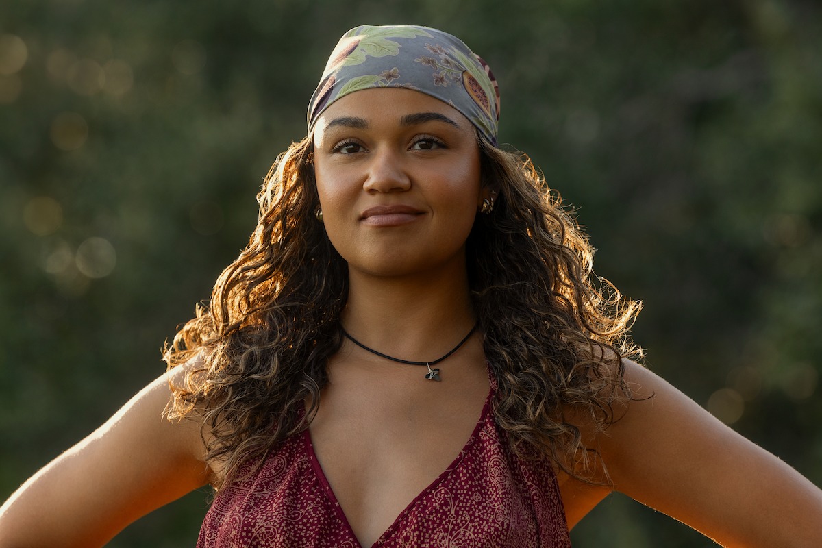 Woman with curly hair and headscarf stands outdoors with hands on hips, wearing a patterned sleeveless top and necklace, in natural light with green blurred background. Confident expression.