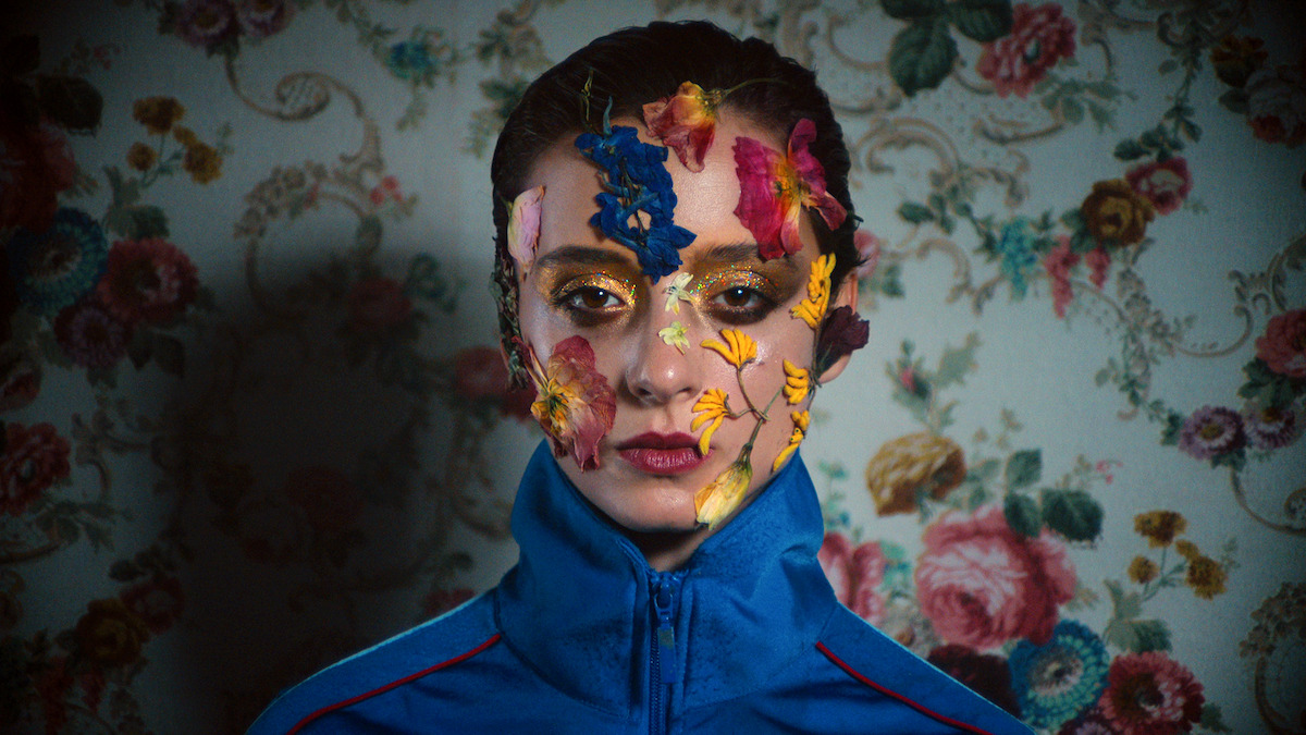 Miu (Angela Bundalovic) in Copenhagen Cowboy (2022). She wears a blue top and butterflies on her face. Against a wall-papered backdrop.