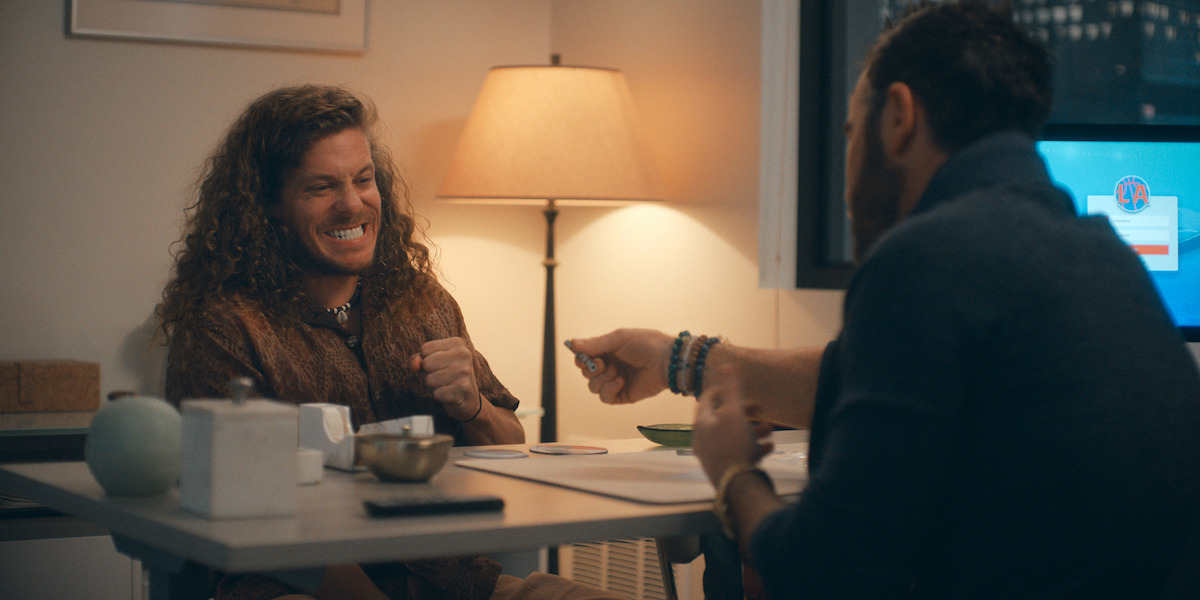 Two people sit across from each other at a small table in a warmly lit room, one grinning and clenching fists, the other handing something over, suggesting a friendly or intense conversation or negotiation. A lamp and computer are in the background.