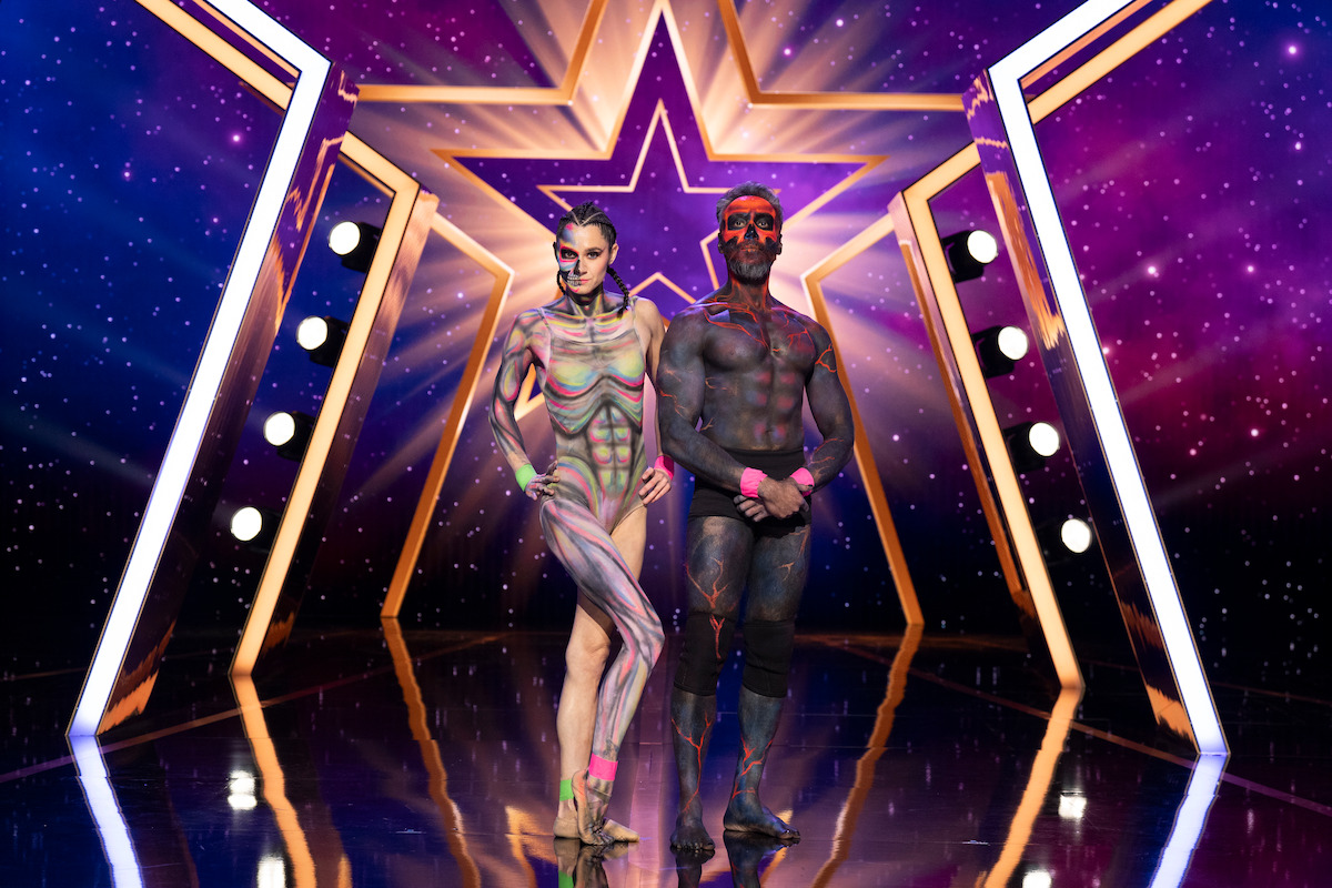 Duo Vespertillio with intricate body paint stand on a shiny stage with a star backdrop, surrounded by bright lights and purple cosmic decorations.
