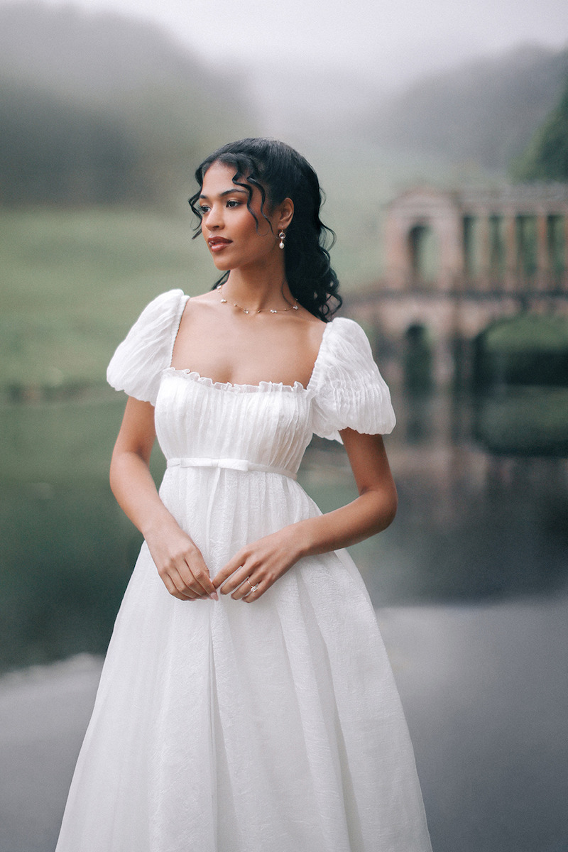 Woman in sleeved bridal dress looks off into distance with a small body of water behind her.