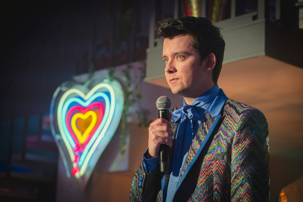 Man in colorful jacket with blue bowtie holding microphone, standing indoors in front of a glowing neon heart sign, under soft, dramatic lighting and festive decor.