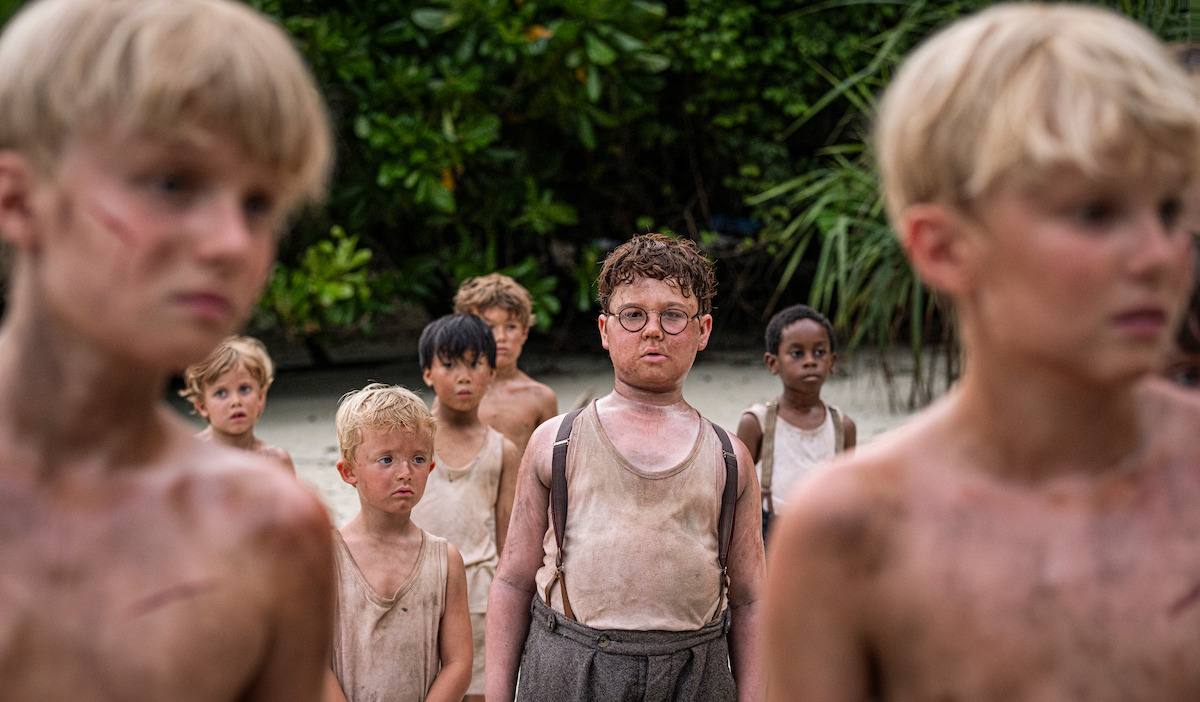 A group of young boys with dirty faces and clothes stand on a sandy ground in a dense jungle, appearing tense and serious, likely stranded or lost in the wilderness.
