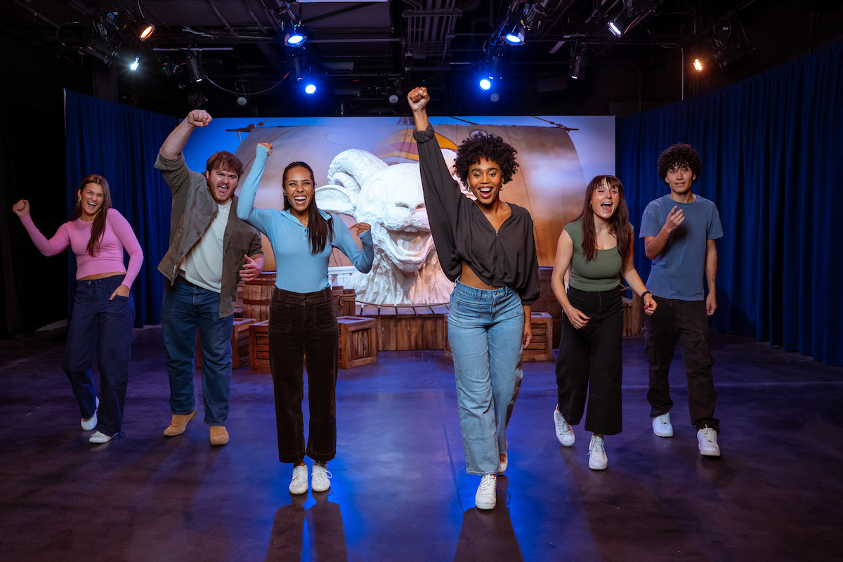 Six people pose enthusiastically on a stage with dramatic lighting, blue curtains, and a theatrical set featuring a large cartoon face in the background, creating a lively and energetic performance atmosphere.