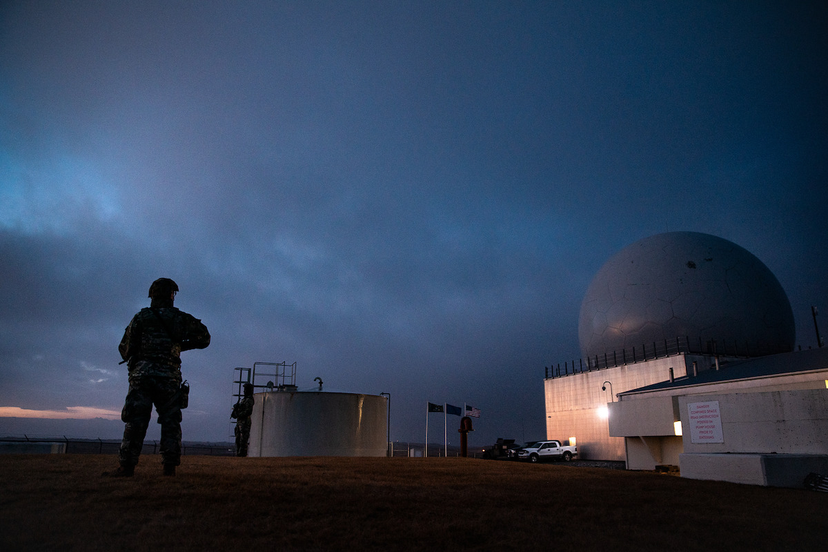 Silhouetted soldier stands guard outside a large radar dome facility at dusk, with flags and vehicles nearby under a dramatic, dimly lit sky, evoking a serious, guarded atmosphere.