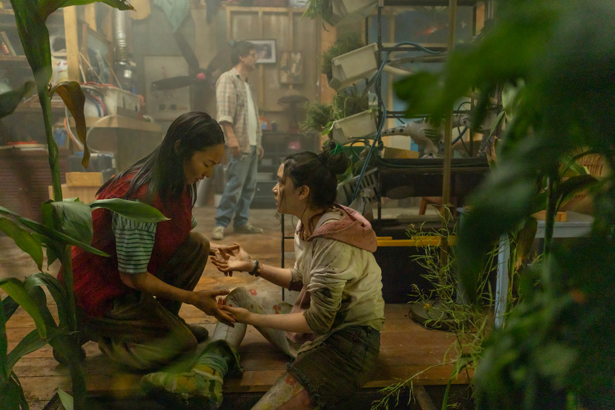 Two women sit on a wooden floor in a cluttered greenhouse, one tending to the other's injured leg, while a man stands in the background among plants, tools, and equipment. The atmosphere is tense and dramatic.