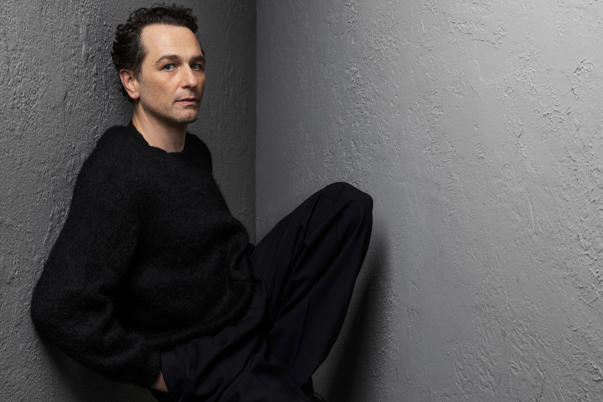 Man in dark clothing sits against the corner of a grey textured wall, looking thoughtfully at the camera in a minimal, moody setting.