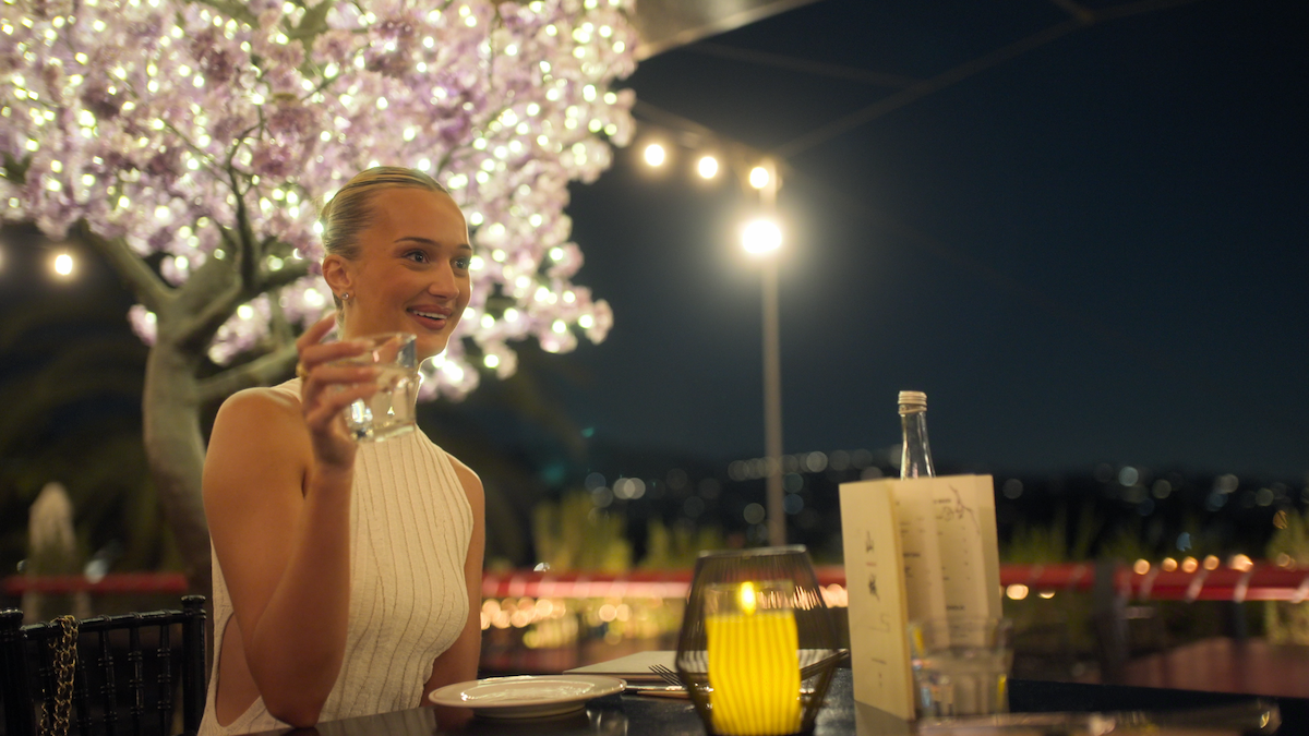 Woman in a white dress sitting at an outdoor restaurant at night, holding a glass, with decorative lights, a glowing tree, menu, candle, and city lights in the background.