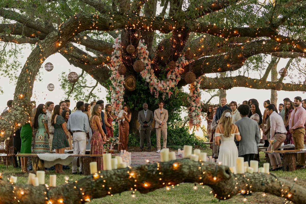 Outdoor wedding ceremony under a large tree decorated with flowers and string lights, with guests gathered around the couple. Candles on tree branches create a warm, romantic setting in a natural environment.